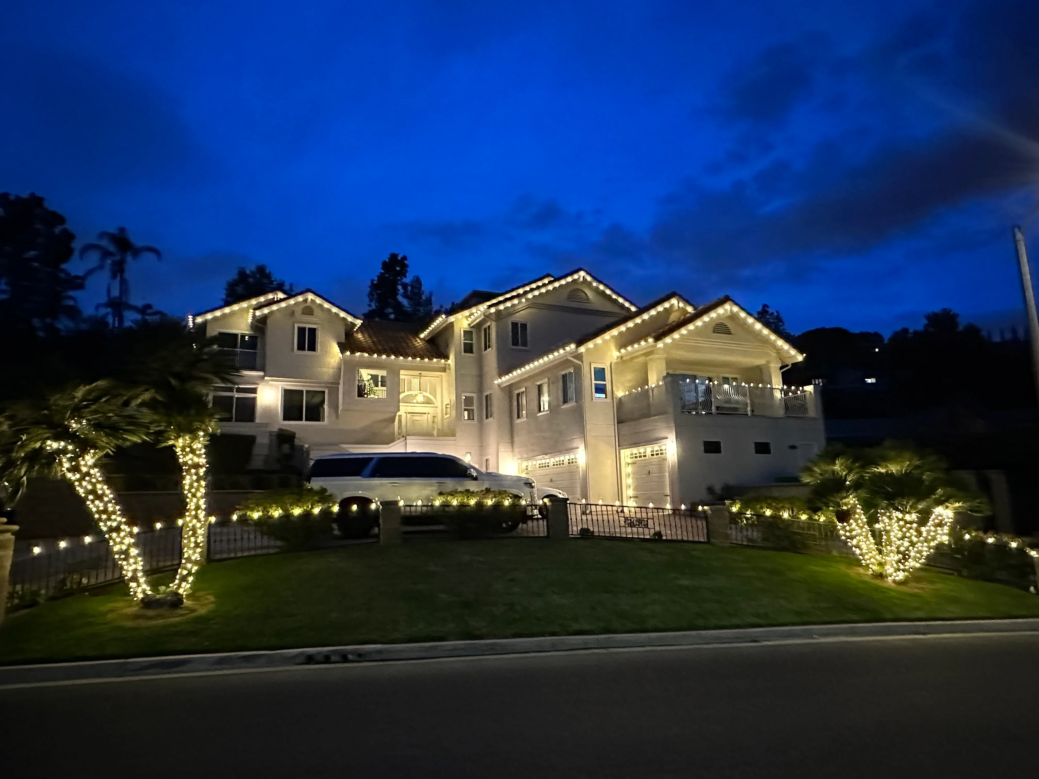Large white Mediterranean estate at dusk with warm-white Christmas C9 lights traced along every gable, palms wrapped warm-white flanking the entry